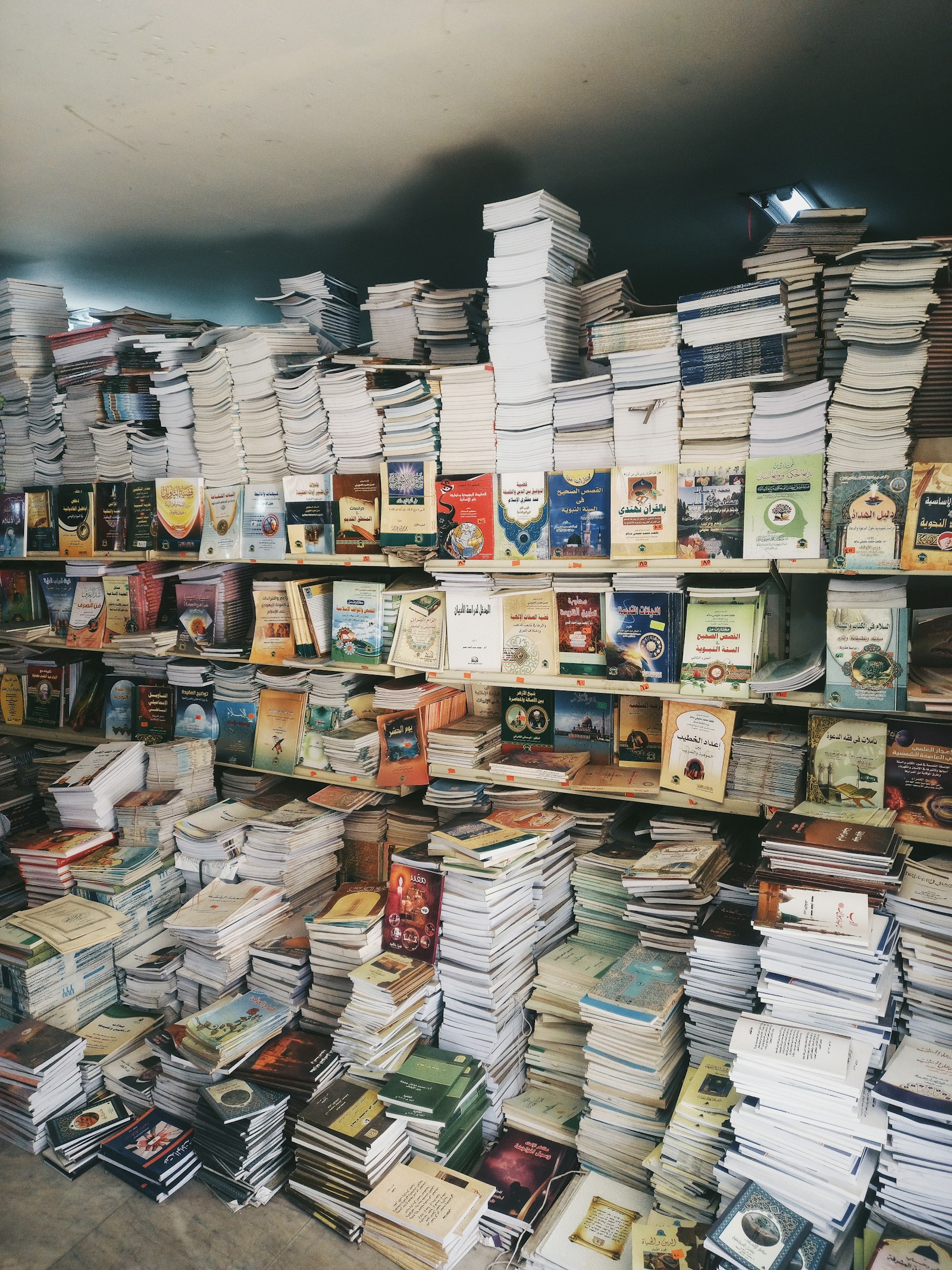 Towering, chaotic stacks of Arabic-language books spilling from overcrowded shelves in a dimly lit bookshop — a sea of titles too vast to navigate without a guide.