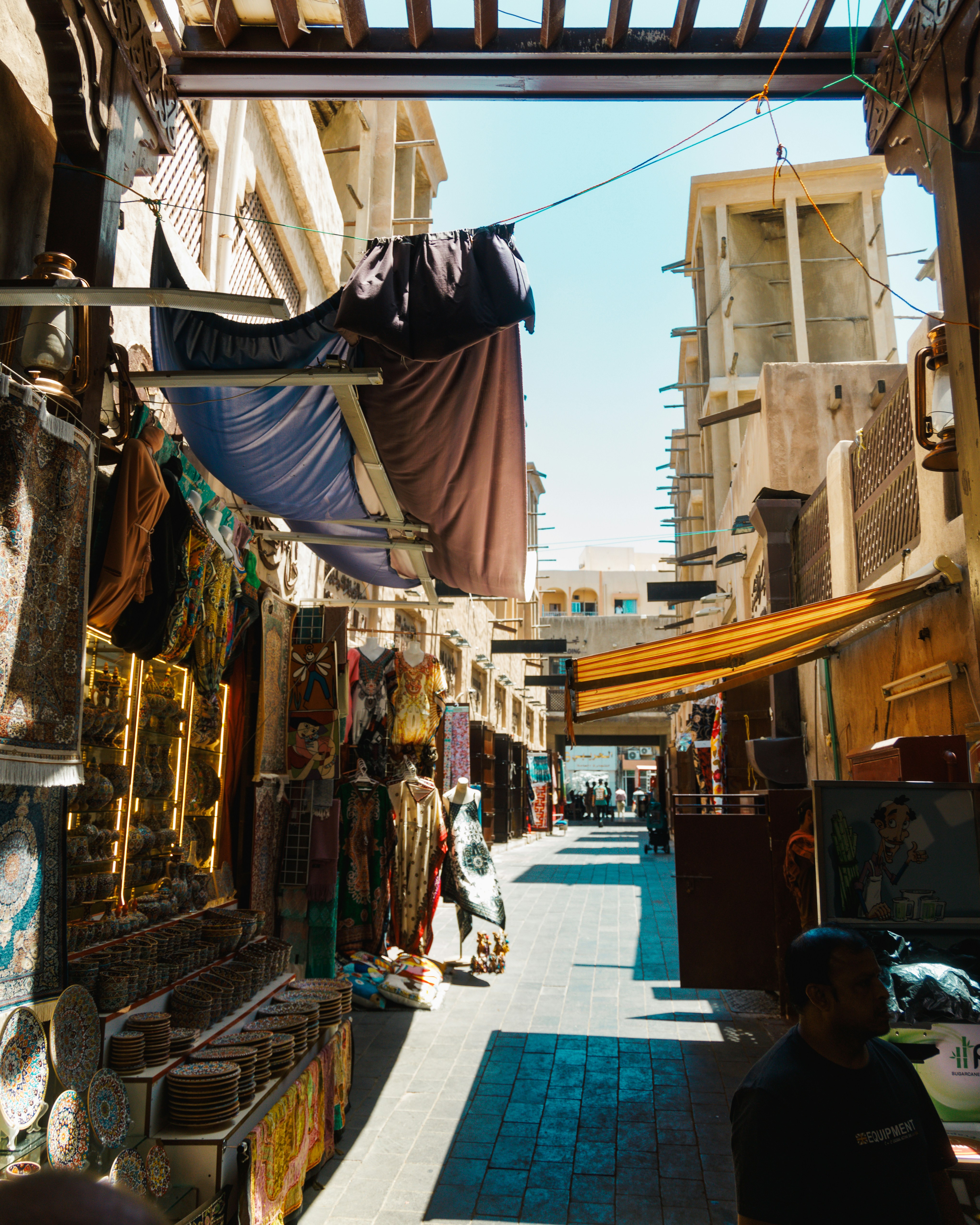 Sunlit alleyway of the Deira Souk in Dubai — textiles, ceramics and traditional wares displayed along narrow stalls with wind towers visible in the distance.