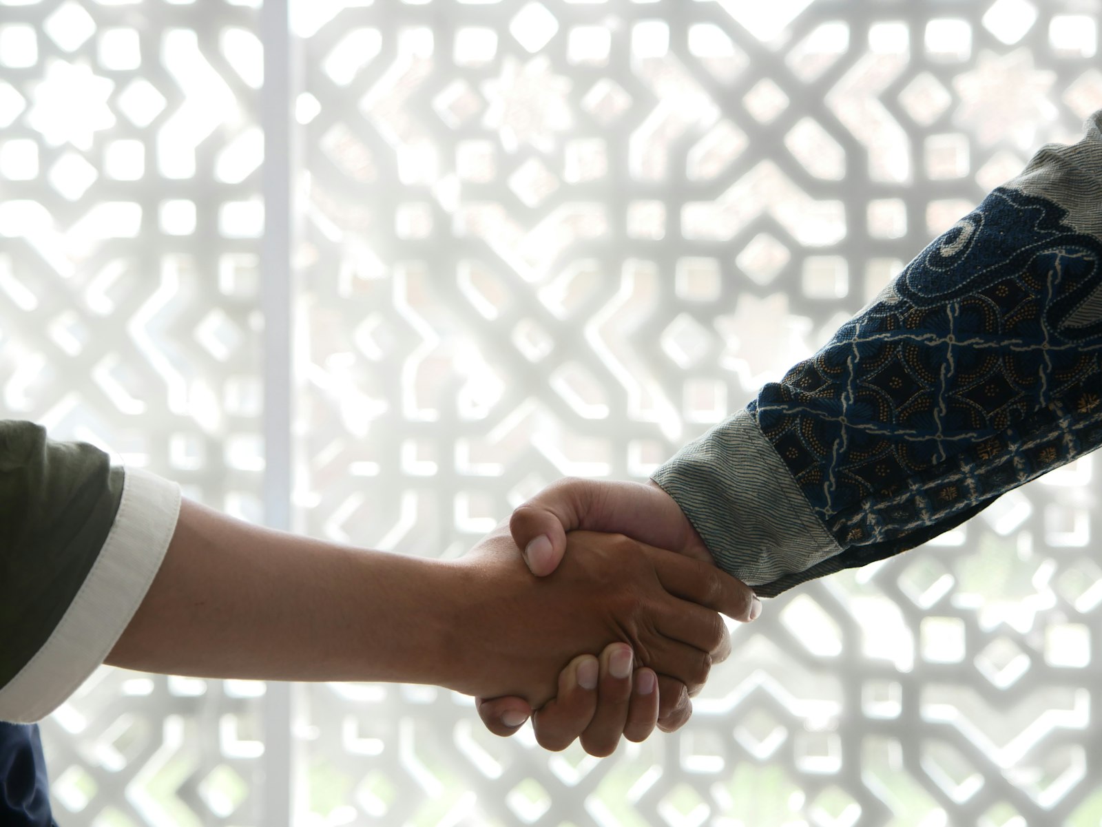 Two hands meet in a firm handshake — one in a green sleeve, the other in a patterned batik shirt — silhouetted against an Islamic geometric lattice screen. A simple gesture of trust and partnership across cultures.