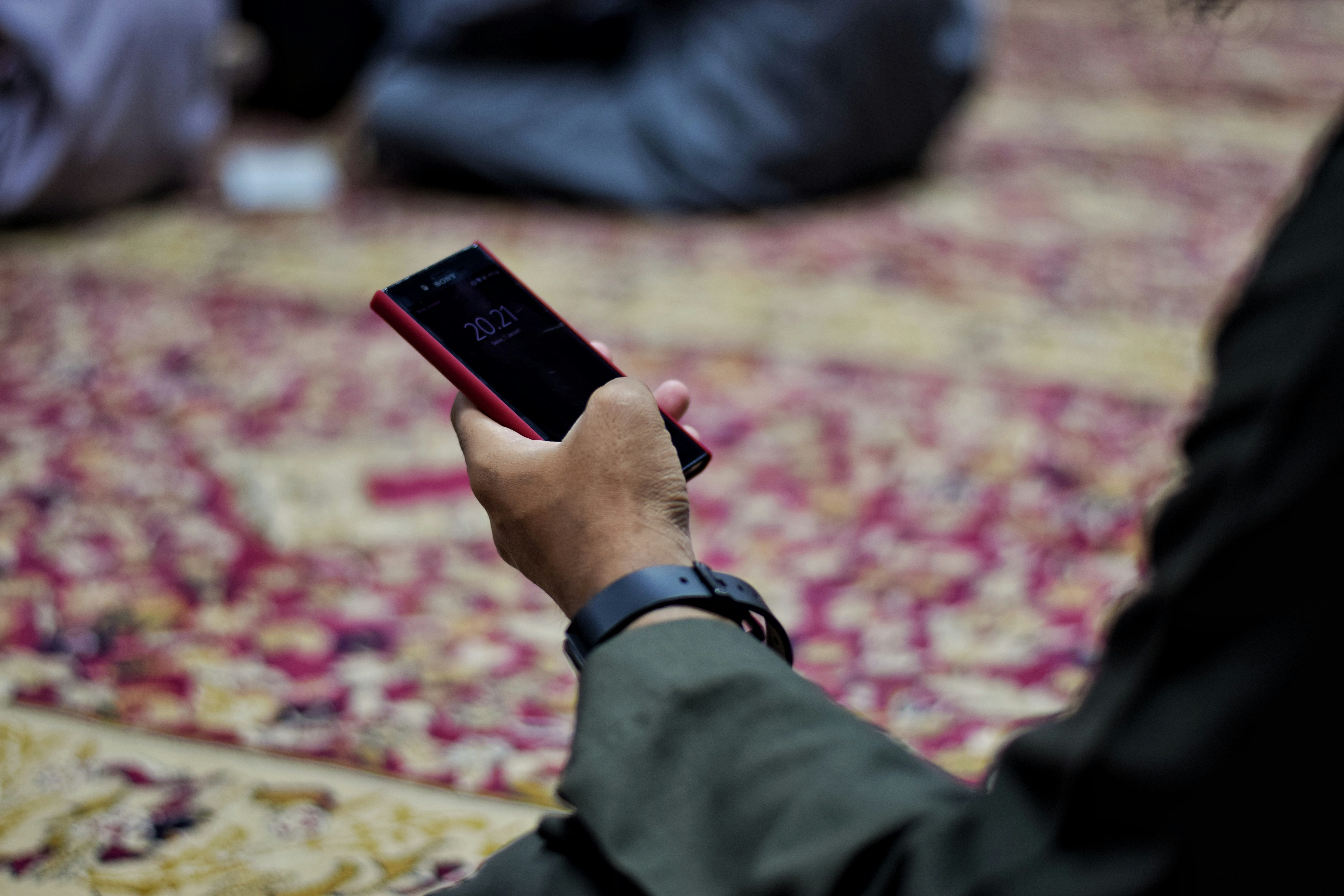 A hand holds a red smartphone displaying the time 20:21 while seated on a patterned prayer rug — a moment of quiet scrolling between prayers.